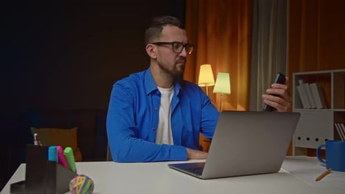 Man Using Laptop and Smartphone and Sitting at Table in Home Office Young Freelancer Looks at
