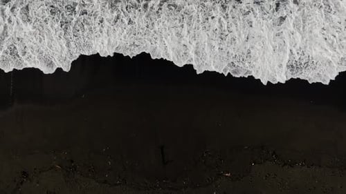 Aerial View of Black Sand Volcanic Beach with Crashing Waves - Top down long drone shot. Serene and