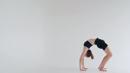 Mujer flexible realizando una pose de rueda con una sola pierna en un estudio blanco
