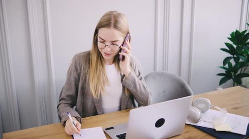 Young Woman Multitasking with Phone and Laptop at Elegant Work Desk in Modern Office Setting