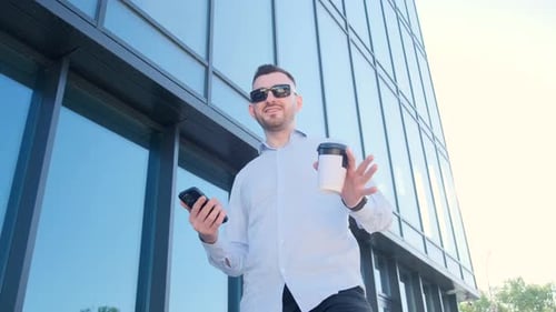 A Handsome Man is Standing Outdoors Near a Skyscraper Drinking Coffee and Talking to the Camera