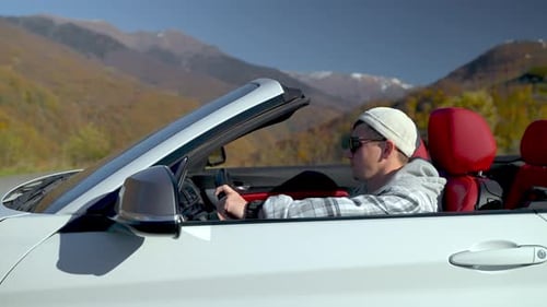 Guy Enjoying Life Sitting in Convertible White Car Among Mountains Side View