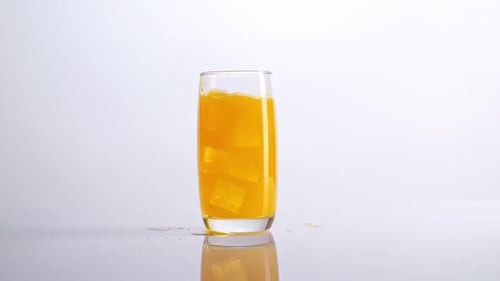 Ice Cubes Falling In A Glass Of Orange Juice On The Light White Screen Background In The Studio