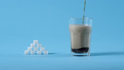 Cola Soda Drink Pouring in Glass and Stack of Lump Sugar on Blue Background