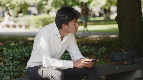 Handsome businessman sitting on bench in park and surfing web, handheld view