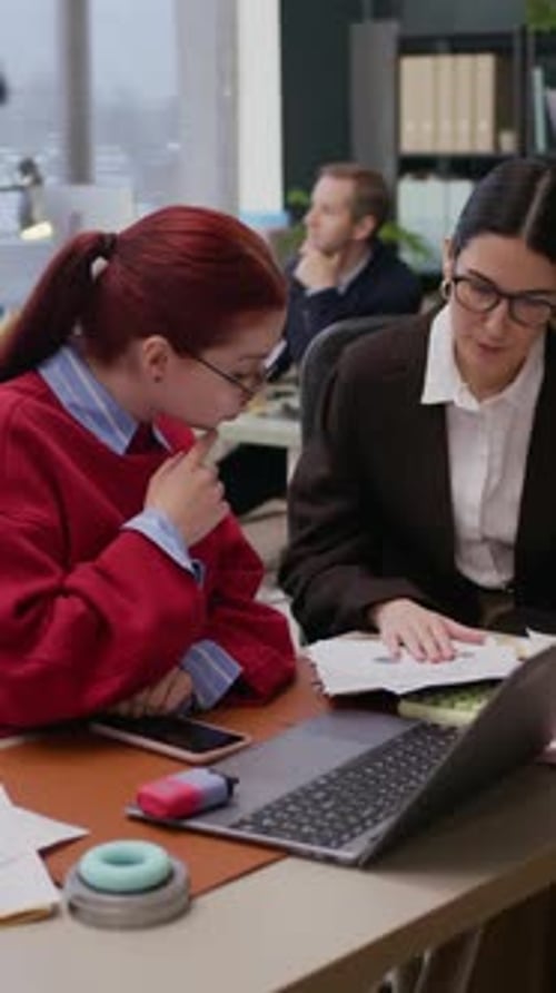 Female Colleagues Discussing Tasks at Office Desk with Papers and Laptop