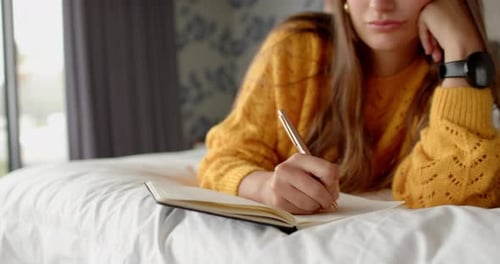 Writing in journal, woman in yellow sweater relaxing on bed at home