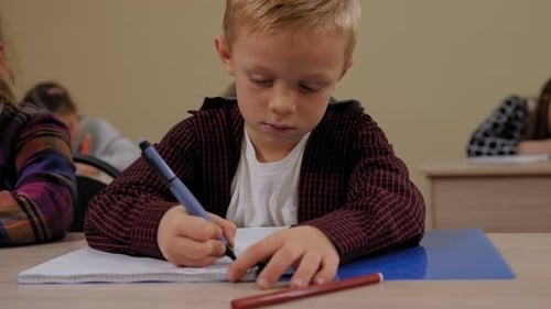 Closeup of a Little Boy Sits at a Desk in School and Writes in a Notebook