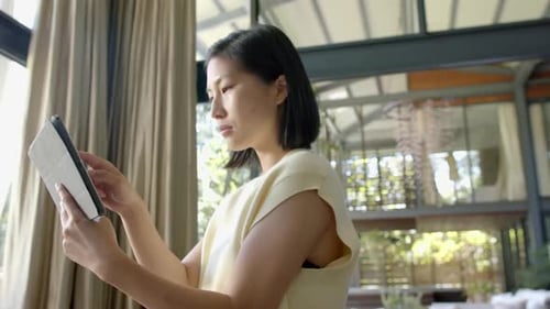 Happy asian woman standing and using tablet in sunny living room, slow motion