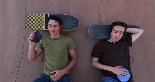 Two Young Men Relaxing on Skateboards Listening to Music From Above