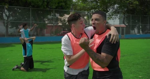 Latin and White teammates celebrate goal on urban soccer field, running with arms in air, pumping