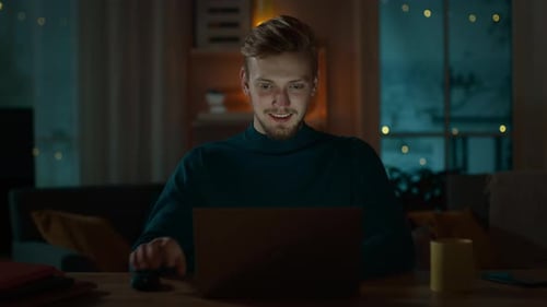 Handsome Smiling Man Works on a Laptop while Sitting at His Desk at Home. Portrait of a Young Freel