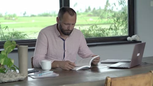 Man Reading Book at Wooden Desk with Laptop