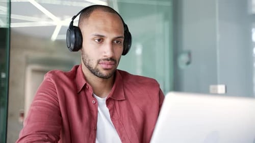 Focused businessman or IT specialist in wireless headphones works on a laptop sitting at workplace