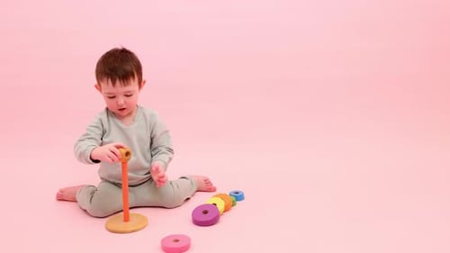 Fröhliches Kleinkind spielt mit einer Pyramide auf einem studio-rosa Hintergrund. Junge legt Holzringe an