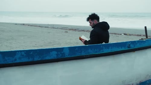 Man Reading A Book Sitting On The Beach In Winter Near A Boat
