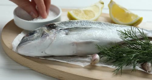 Woman adding salt onto fresh raw dourade at white wooden table, closeup. Fish and seafood delicacies