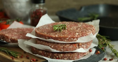 Putting uncooked meat patty onto grill pan, focus on stack of patties with rosemary on table