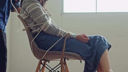 Cinematic side view of hostage victim bound to chair by unidentified captor in balaclava