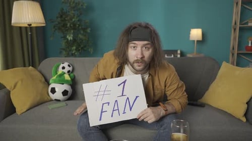 A Man is Sitting on a Couch He is Watching a Soccer Game on TV and Holding a Fan Sign He Shows