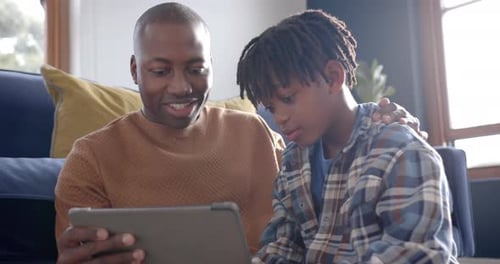 Happy african american father and son sitting on floor and using tablet at home, slow motion