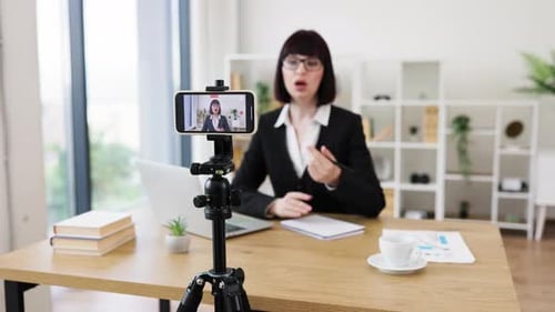 Businesswoman Recording Blog Seated at Office Desk with Camera and Laptop