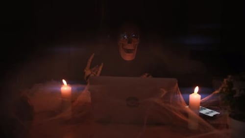 Death character in mystical fog move bone jaw while sitting at table in laptop among cobwebs