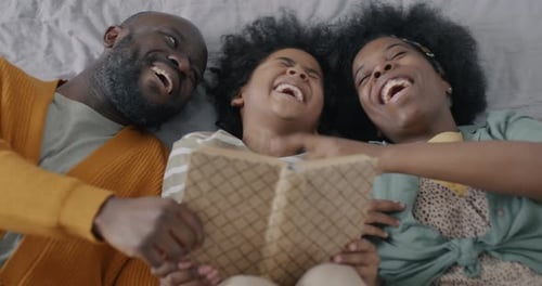 Top View of Joyful African American Family Mom Dad and Child Reading Book and Laughing Lying in Bed