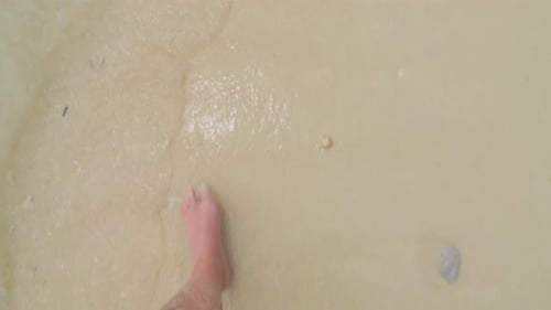 A male's bare feet walking on a sandy beach, with gentle sea waves lapping at his ankles.