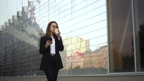 A Young Respectable Woman Walks Down the Street with a Smartphone and Drinks Coffee