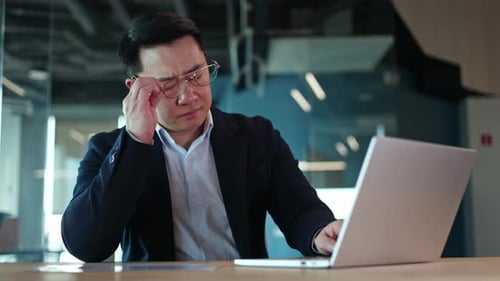 Male Office Worker Having Headache and Taking Off Glasses While Typing on Laptop