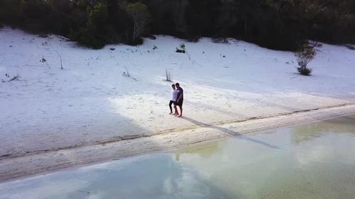 Drone footage of a couple walking down a remote beach with beautiful white sand while holding hands.