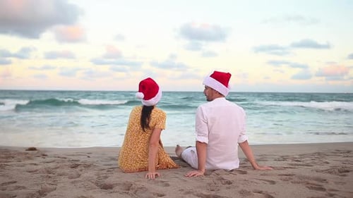 Young Couple in Red Santa Hats Sitting on the Beach on Christmas