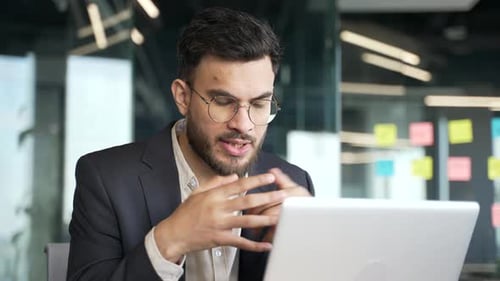 Confident businessman in a formal suit talking on a video call using a laptop at workplace in office