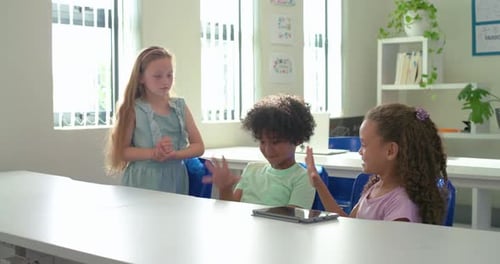 Diverse kids at school table, girl in blue dress approaching, initiating playful game, using tablet
