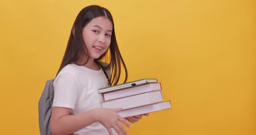 Cute school girl is carrying a school bag and holding a textbook on a yellow background.