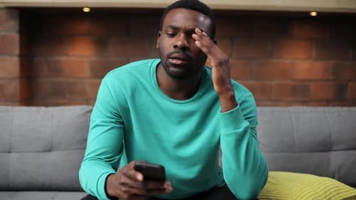 Portrait of Focused Likable African Guy in Mint Sweater Holding Smartphone in Hand and Looking