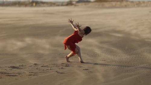 Graceful Smiling Young Woman in Red Evening Dress Dancing on Sandy Beach in the Wind