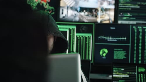 Hooded Computer Hacker Coding on Computer While Sitting at Office Full of Monitor Screens