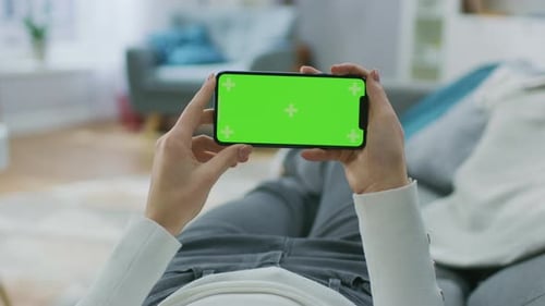 Young Woman at Home Lying on a Couch using with Green Mock-up Screen Smartphone in Horizontal Lands