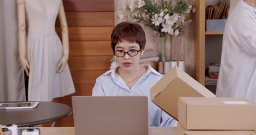 Adult asian daughter holding cardboard box beside laptop boutique fashion studio teamwork e-commerce