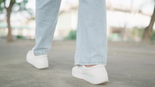 A rear view video follows a woman's feet walking down a street on a sunny day. A woman wearing sneak