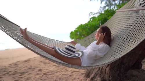 Asian woman swinging in the hammock while facing the calm sea. Carefree girl enjoying the sunset on