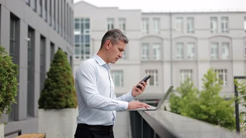 Man Talks on Phone While Using Laptop Outdoors