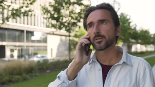 A Handsome Caucasian Man Argues on a Smartphone As He Walks Down the Street in an Urban Area