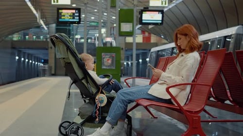 Young mother waits at subway station with baby in stroller while using phone