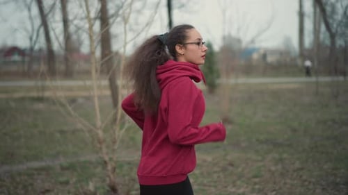 Young Woman Running Side Profile Along Woodland Path In Red Hoodie And Glasses Focused Tempo Pacing