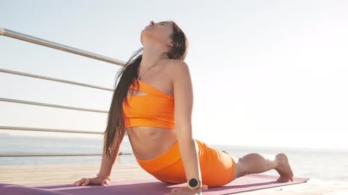 Young Woman in Orange Sports Wear Practicing Yoga on the Beach Above Sea at Amazing Sunrise
