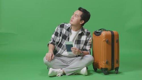Full Body Of Asian Male Traveler With Luggage And Looking Around While Sitting In The Green Screen