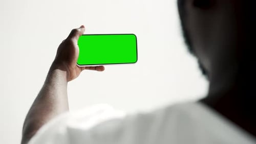 Young african american man looks at green screen smartphone in hand indoors